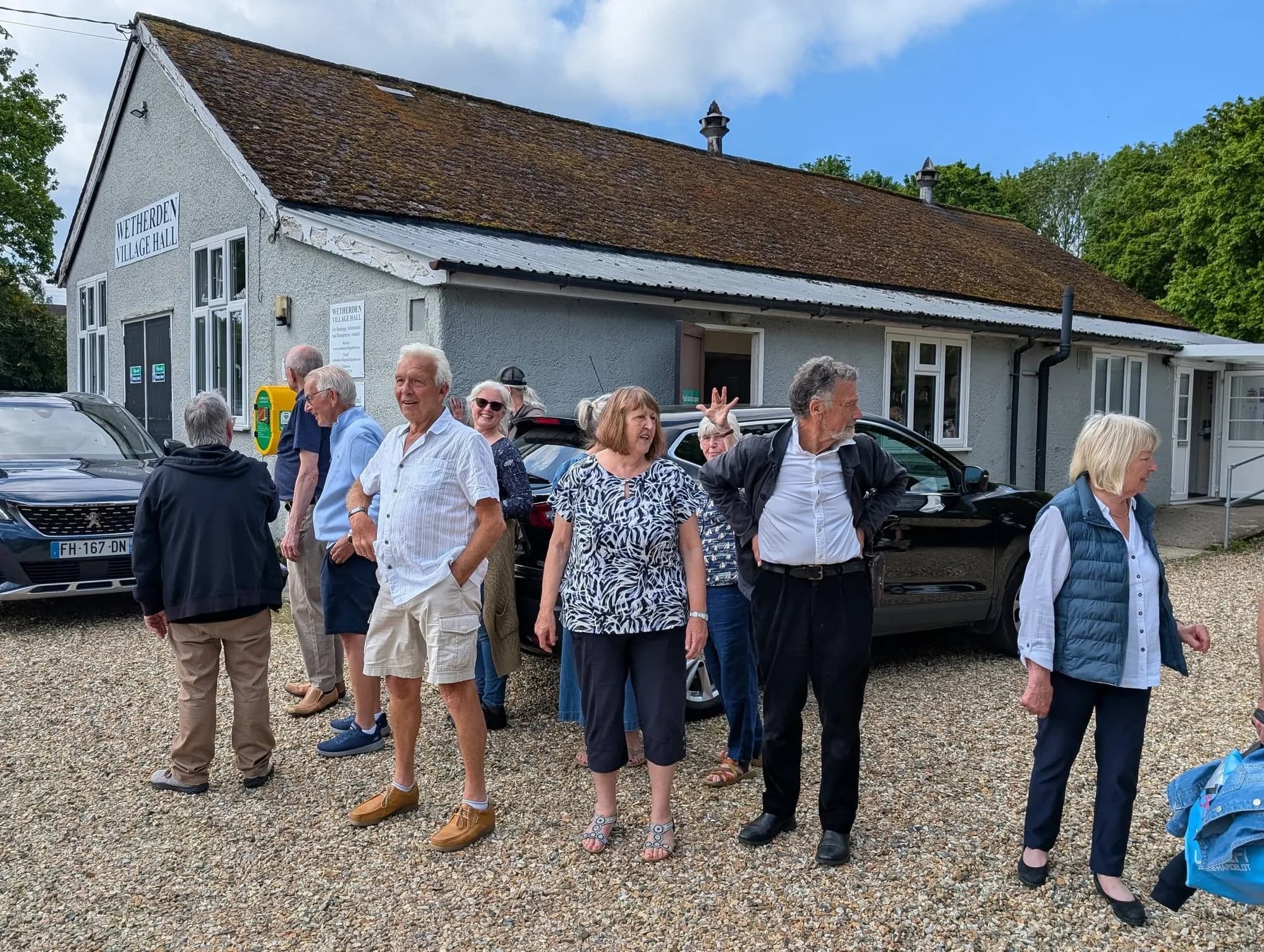 A group of older adults mingles and chats on the gravel car park outside Wetherden Village Hall on a sunny day, with parked cars visible behind them, during the HAWTA French visit farewell brunch.